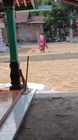 A person wearing a pink floral dress and a green head covering sweeps a large area covered in light brown material. The person is positioned in the middle of the frame, facing away from the camera. The area being swept is outdoors and appears to be a drying area, possibly for grains or agricultural produce. In the background, there are buildings, trees, and flags. A motorcycle and a few sacks are also visible in the distance. A broom is leaning against a pillar in the foreground to the left of the frame. The ground in the immediate foreground is a mixture of dirt and tiles.