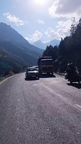 A video shows a road bordered by a mountain and trees. A series of vehicles are seen driving on the road. The sky is blue with clouds, and the sun is visible. The video appears to be filmed from a moving vehicle.