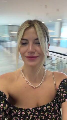 A young woman with blonde hair and a floral dress is recording a video of herself. She is wearing a necklace and earrings. The background appears to be an indoor shopping mall or store, with blurred displays and escalators visible. The woman smiles and makes various facial expressions, looking directly at the camera. The video has a dynamic, selfie-style feel, likely captured while walking or moving.