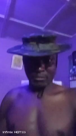 A shirtless man wearing a camouflage bucket hat is standing indoors, looking towards the camera. The room is dimly lit with a dominant purple or blue hue, suggesting artificial lighting. He slowly turns his head from his right to his left and back again throughout the video. In the background, to the right of the man, there is a light-colored appliance, possibly a refrigerator, with the word "BOSCH" visible on its side. To the left, a framed item or picture is hanging on the wall. "Infinix HOT 50i" is visible as a watermark in the bottom left corner of the video.