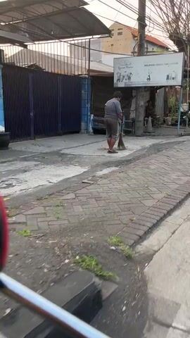 A person is sweeping the ground near a building with a blue metal gate. The person is wearing a gray t-shirt and shorts. There is a sign on a pole near the person. The ground is paved with bricks and asphalt. There are some plants growing in the cracks of the pavement. The scene appears to be an outdoor area in a city.