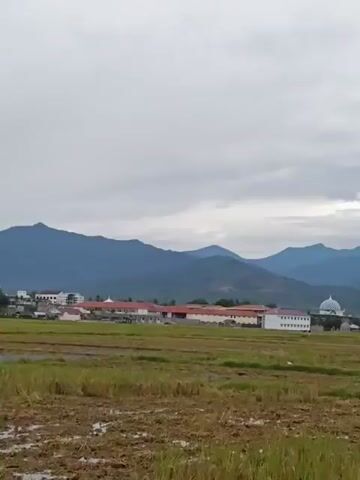 The video pans across a rural landscape with a harvested rice field in the foreground. In the mid-ground, several modern buildings are visible, including a domed structure resembling a mosque, and a long white building with a red roof. Further back, a range of blue-green mountains forms the backdrop under a cloudy sky. A bird flies across the sky at the 0:02 mark.