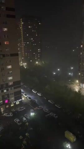 The video shows a nighttime scene in an urban environment with heavy rain. Two tall apartment buildings flank the view. In the foreground, a parking lot is visible with several cars parked. A line of trees separates the parking lot from a road further in the distance, which is illuminated by streetlights. The rain is so heavy that visibility is reduced, creating a foggy or misty effect. Some windows in the apartment buildings are lit up, indicating occupancy. The focus is on the heavy rainfall and the urban landscape at night. There are no visible people in the scene.