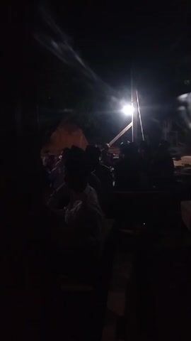 A group of people are gathered outdoors at night, illuminated by a single bright light source. The figures are largely in silhouette against the light, making individual identification difficult. Some individuals appear to be wearing traditional clothing and headwear. The environment seems to be rustic, with some structures and possibly building materials visible in the background. The atmosphere is dark and somewhat obscured, suggesting a communal gathering or event taking place after sunset.