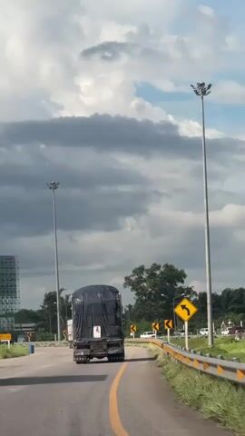 The video shows a view from inside a vehicle driving on a road. In front of the vehicle are two trucks. The truck in the left lane is white with a tan cargo container. The truck in the right lane is dark colored, possibly black, and its cargo container is covered with a dark tarp. Both trucks are moving away from the camera. The road has a yellow dividing line and a yellow barrier on the right side. There are tall light poles on both sides of the road. The sky is cloudy with patches of blue sky. There are traffic signs visible, including a yellow diamond-shaped sign with a black arrow indicating a curve to the left. The environment appears to be rural or semi-rural, with trees and vegetation visible in the background.