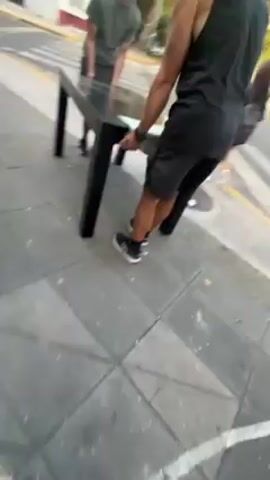Two men are carrying a black rectangular table across a crosswalk. The table has four legs. The men are dressed casually in shorts and t-shirts. One man is Black and the other has brown hair and a beard. They are walking on a paved sidewalk in an urban environment. There are cars parked on the street and buildings in the background. A woman crosses the crosswalk separately. The camera then pans to follow the sidewalk, showing a wall with graffiti and more trees and parked cars. The weather appears to be sunny.