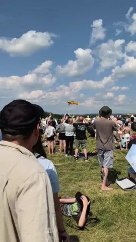 A yellow airplane flies through a clear blue sky dotted with white clouds. The plane is emitting a white smoke trail. Below, a crowd of people stands on a grassy field, watching the airplane. Some people are holding umbrellas. There are tents set up in the field. The overall scene suggests an outdoor event, possibly an airshow.