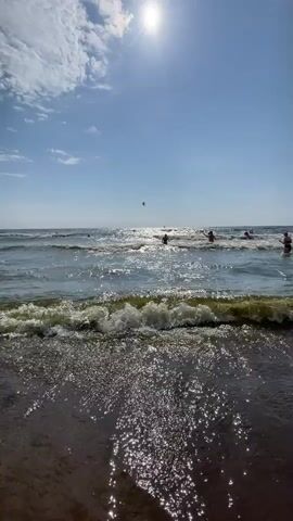 The video shows a beach scene with the sun shining brightly in a blue sky with some clouds. The ocean is calm with gentle waves breaking on the shore. Several people are in the water, some swimming and others standing. The camera is at a low angle, close to the water, looking out towards the ocean. The waves are creating foam and spray as they come in. In the distance, there appear to be other people on the beach or in the water. The overall atmosphere is one of a pleasant summer day at the beach.