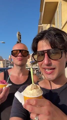 The video features two individuals enjoying gelato on a sunny day. The person in the foreground, wearing sunglasses and a black t-shirt, smiles at the camera while holding a cup of light-colored gelato with a green spoon. Behind them, to the left, another person with a shaved head, also wearing sunglasses and a black tank top, holds a cup of pink gelato and a green spoon. The background reveals a picturesque European setting with buildings, a street, and a marina. In the middle ground, several large yachts are docked, indicating a harbor or waterfront. A black car is parked nearby, and a red carpet-like surface is visible in the lower left corner. A sign with the word "TAXI" in white letters on a red background is also present. The clear blue sky suggests fair weather and daytime. The overall mood appears relaxed and cheerful, with both individuals seeming to enjoy their treats and the surroundings. The time of day is likely afternoon, given the bright sunlight and clear skies. The location context is identified as Saint-Tropez, France, which aligns with the presence of luxury yachts and the charming architecture.