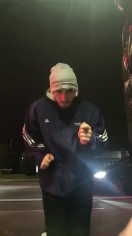 A young man wearing a light grey beanie, a blue Adidas tracksuit jacket, and black pants is dancing on a street at night. His hands move rhythmically. The background features a dark sky, streetlights, a road with white lines, parked bicycles, a building with illuminated signage, and a black car with its headlights on. The overall environment is urban and dark.