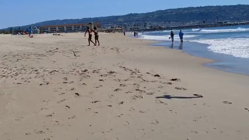 The video shows a beach scene on a sunny day. The camera is angled slightly upward, focusing on the beach and the ocean. There are several people on the beach, some walking along the shoreline and others standing near the water's edge.  A couple is walking along the water's edge, a person in dark clothing is standing in the foreground, and several other people are seen further down the beach.  Seagulls are flying overhead. The ocean has small waves, and in the distance, there is a landmass with buildings visible. The sky is clear and blue.