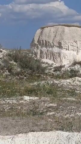 The video shows a moving shot of a dry, arid landscape. The foreground consists of light-colored, rocky terrain with sparse, dry vegetation. Further back, there are slopes covered in more vegetation and some white, rocky formations. The sky above is blue with scattered clouds. The camera appears to be moving horizontally, panning across the scene.
