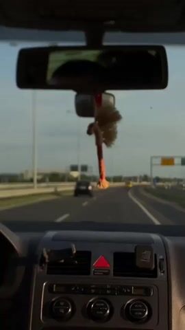 The video shows a view from inside a car driving on a highway. The road ahead is visible through the windshield, with other cars in the distance. The car's dashboard is in the foreground, and a decorative object hangs from the rearview mirror. The sky is visible in the background, with some clouds. The camera appears still inside the vehicle. The main action consists of the car moving along the road.