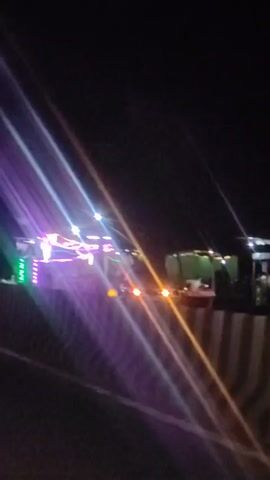 The video shows a nighttime scene from a moving vehicle. The foreground is dark, with a concrete barrier on the right side of the road. In the distance, a truck is decorated with an abundance of colorful lights, including purple, pink, green, and white. The lights are strung across the truck and appear to be part of a celebration or event. Several people are visible near the truck, silhouetted against the bright lights. The overall atmosphere suggests a festive or celebratory gathering occurring at night on or near a road.