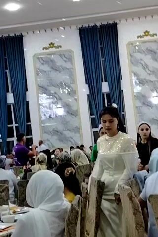 The video shows a large indoor event, possibly a wedding reception or banquet, with many people seated at tables. The room is decorated with blue curtains and large marble-patterned wall panels. A prominent chandelier hangs from the ceiling, and there are smaller lights scattered across the ceiling. In the foreground, a young woman in a white dress with beaded embellishments is standing and looking towards the camera. In the middle ground, several people are visible, some seated at tables, others standing. A man in a purple shirt is standing on the left, holding up a phone, seemingly recording. In the background, more guests are seated, and a group of musicians appears to be performing. There are also several bottles of drinks and food on the tables. The overall impression is of a festive gathering.