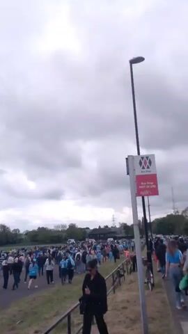 A large crowd of people is walking on a road and a grassy area next to it. Most of the people seem to be wearing blue or light blue clothes. There are some trees and lampposts along the road. There is also a bus stop sign that says "Bus Stop NOT IN USE". The sky is cloudy and overcast.