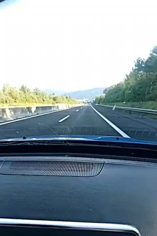 This is a first-person perspective video, likely from inside a vehicle, looking forward through the windshield. The view is of a highway with multiple lanes. On the left side of the road, there is a concrete barrier and green trees beyond it. On the right side, there is also vegetation. The sky is bright and cloudy. The dashboard of the vehicle is visible at the bottom of the frame, with a speaker grille. The road appears to be in good condition, with clear lane markings. There are other vehicles visible in the distance on the highway. The video appears to be shot during the daytime.