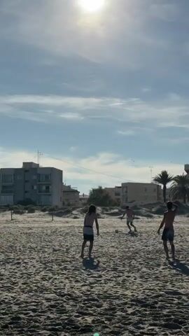 Three shirtless people are playing soccer on a sandy beach. One person falls onto the sand after attempting to kick the soccer ball. Buildings and palm trees are in the background. The sky is blue with some clouds. The sun is visible.