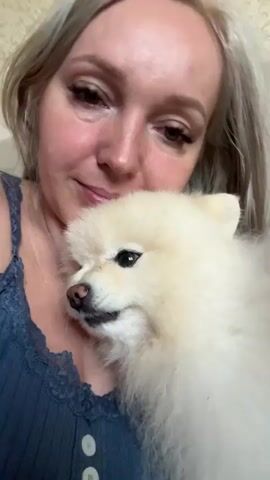 A woman with blonde hair is holding a fluffy white dog up to her face. The dog is looking to the side with its mouth slightly open, revealing its teeth. The woman is looking down at the dog and appears to be smiling. She is wearing a blue tank top with lace trim. The background is blurred and appears to be a wall with a textured pattern.