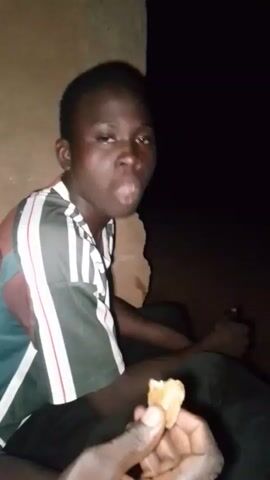 A young person with dark skin and short hair is sitting down, leaning against a wall. They are wearing a t-shirt with vertical stripes in green, white, and red. The person's right hand is extended and appears to be holding a small, light-colored object. The background is dark and indistinct, suggesting it is nighttime or a poorly lit area. The person is looking towards the right side of the frame.