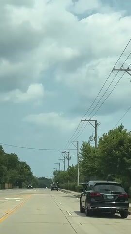 This is a video taken from the perspective of a driver on a paved road. The sky is partly cloudy and a light blue. The road has two lanes in each direction, separated by a double yellow line. There are trees and power lines lining the right side of the road. Several vehicles are visible on the road, including a black SUV in the foreground on the right, and a few cars further ahead. The SUV has a license plate that is partially visible. The video appears to be taken during the daytime.