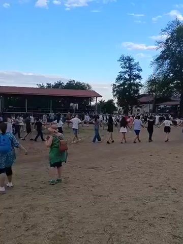 A group of people are dancing in a circle holding hands in an outdoor dirt area. There is a covered pavilion with a red roof in the background, and trees and buildings beyond that. The sky is blue with some clouds. The people are dressed in casual clothing.