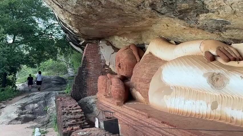 The camera pans from a view of a large, partially reconstructed, reclining Buddha statue built into a rock wall to a view from atop a hill overlooking a dense forest. Two people walk along a rocky path, and a group of about 10 people are standing at the edge of a lookout point, observing the landscape.  The terrain is rocky and surrounded by lush green trees and shrubs. In the distance, rolling hills are visible under a cloudy blue sky.  A green lamp post stands in the foreground.
