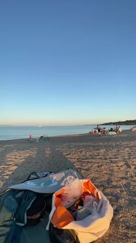 The video opens with a close-up of beach gear in the foreground, including a dark bag, a turquoise backpack, and a white bag overflowing with items such as an orange towel or shirt and plastic bags. The camera then pans out to reveal a sandy and pebbly beach with shadows cast across it. In the middle ground, several people are gathered, some sitting and others standing, appearing to be enjoying the beach. Further in the distance, the calm sea stretches to the horizon under a clear, pale blue sky. A single sailboat is visible on the water, and a faint outline of land can be seen on the horizon. A few small white buoys are dotted across the sea. The overall scene suggests a peaceful late afternoon or early evening at the beach.