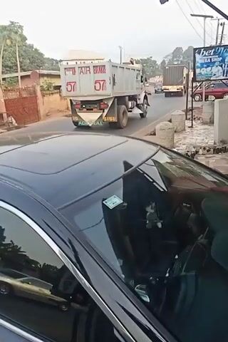 The video shows a street scene, apparently shot from inside a car. In the background, trucks are driving down the road. A sign for “bet9ja MEGA SHOP” is visible. The camera shifts to a person standing outside a building painted red and white. The person is wearing a yellow patterned shirt, dark shorts, a light-colored hat, and dark sandals. They are looking at a cell phone. The camera pans back to the street showing more vehicles. A car horn is heard.