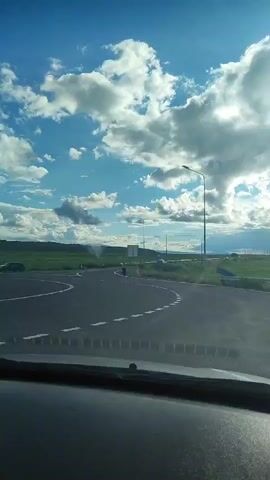 The video shows a view from inside a vehicle, looking out at a roundabout on a clear day with a blue sky and white clouds. The camera is positioned behind the dashboard. The roundabout has white dashed lines marking its lanes. A dark-colored car is driving on the roundabout, and it is the primary focus of the scene. The surrounding environment appears to be rural, with green fields and distant trees visible. There is also a street light visible on the right side of the road. The video appears to be a continuous shot.
