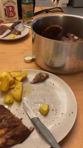 The video shows a messy dining table after a meal. There are dirty plates with leftover food, including roasted potatoes and meat. A large pot filled with meat is visible on the right side of the table. Various food items and utensils are scattered around, suggesting a recently finished meal. The lighting suggests an indoor setting, possibly a home kitchen or dining room. There are no people visible in the frame.
