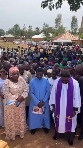 The video depicts an outdoor funeral gathering of a large crowd. The group is gathered around a wooden casket decorated with floral arrangements, presumably containing the body of the deceased. Attendees include both men and women, diverse in age and wearing a variety of clothing. Prominent figures standing near the casket include a woman in a patterned dress, a man in a light blue robe, and a clergy member in white robes with a purple stole. They appear to be officiating or paying respects. The setting is a rural or semi-rural area, with scattered houses and trees visible in the background, suggesting a village or small town location. The crowd is generally somber and attentive.