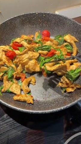 The media file shows a close-up shot of chicken strips, cherry tomatoes, and spinach being stir-fried in a dark-colored pan. The ingredients are being tossed and mixed with a black spatula. The food appears to be seasoned and is glistening, suggesting it is being cooked in oil. Steam is rising from the pan, indicating that the food is hot. The background is out of focus but appears to be a kitchen setting, possibly a stovetop. The lighting suggests it is daytime or a well-lit indoor space. There is no visible text in the media file. The activity taking place is the cooking of a meal. The subjects are food ingredients and cooking utensils. The setting is a kitchen. No people are directly visible, but the presence of the spatula implies someone is cooking.
