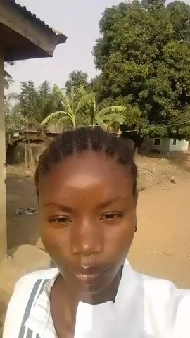 A close-up video shows a young woman with dark skin and braided hair. She is wearing a white shirt and standing outdoors in what appears to be a rural or less developed area. Behind her are buildings, trees, and a dirt ground. The camera is held at eye level, focusing on her face. The lighting is bright, and she appears to be looking directly at the camera. The video seems to have been taken on a clear day.