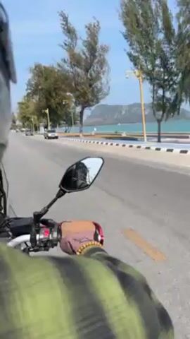 The video shows a person riding a motorcycle along a road, presumably on a sunny day. The rider is wearing a helmet and a green plaid shirt with gloves on their hands. The view is from behind the rider, offering a perspective of the road ahead and the surrounding environment. A mirror on the motorcycle shows the rider's face. The road runs alongside a beach with turquoise water. Trees and street lights line the road, and other vehicles can be seen in the distance. The sound of the wind is prominent.