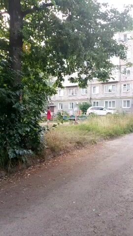 The video shows an outdoor scene with a large tree on the left side. In the background is a multi-story apartment building. In front of the building, there is a small playground area with some equipment. There are two children playing in the playground. One child, wearing a red dress, is standing near a blue swing set. Another child is sitting on the swing set. There is a white car parked in front of the building. The ground in the foreground is dirt and gravel. The overall environment appears to be a residential area.