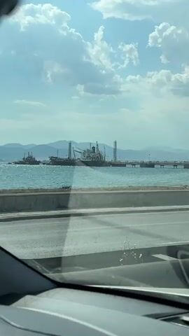 A video shot from inside a car shows a scenic view of a port on a sunny day. The sky is blue with fluffy white clouds. In the distance, large cargo ships and smaller tugboats are visible on the turquoise water. A pier or breakwater extends into the sea, with tall structures that appear to be part of a loading or unloading facility. The shoreline features some greenery and construction materials, including piles of sand. The camera is moving, suggesting the car is in motion.