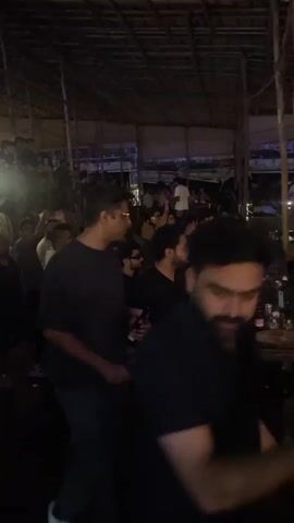 The video shows a dimly lit, crowded indoor space that appears to be a bar or club. The ceiling is constructed of natural materials, possibly bamboo or thatch, supported by wooden beams and poles. The lighting is primarily artificial, with some spotlights and ambient light sources creating a festive atmosphere. A large number of people are visible, many of them standing, talking, and dancing. Some individuals are positioned near a bar area, where bottles and glasses are present. The overall scene suggests a social gathering or event in progress. The camera movement is somewhat shaky, indicating it might be handheld.