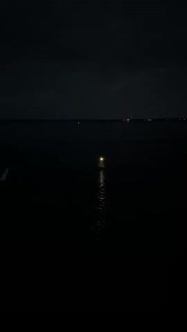 The video shows a night scene of a body of water with a faint reflection of lights in the distance. In the center of the frame, a small lighthouse with a bright light is visible, its reflection creating a shimmering path on the water. The sky is dark and cloudy.