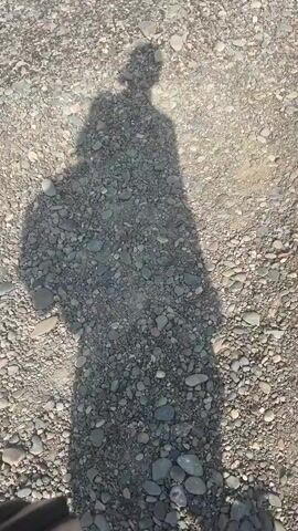 A shadow of a person with a backpack is cast on a gravelly ground. The person appears to be walking. The shadow extends from the top of the frame to the bottom. The environment is outdoors, with sunlight illuminating the scene.