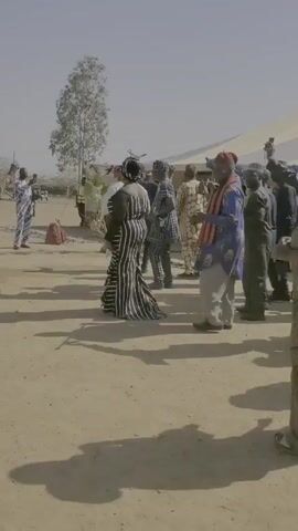 The video shows a group of people gathered outside under a large, light-colored tent. Many of them are dressed in patterned clothing. Some are recording with their phones. They appear to be moving and celebrating together. The ground is dry and dusty. The sky is light blue and mostly clear.
