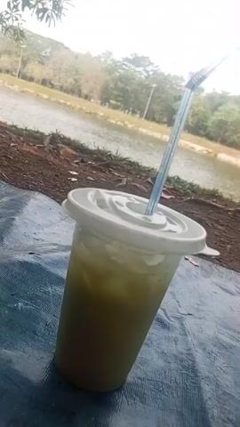 A close-up shot, taken from a slightly low angle and tilted to the right, shows a plastic cup of green liquid with a white lid and a blue and white striped straw inserted into it. The cup is resting on a blue tarp. In the background, a body of water, possibly a lake or pond, is visible, with trees and some structures along its edge. The overall lighting suggests an outdoor setting during the day.