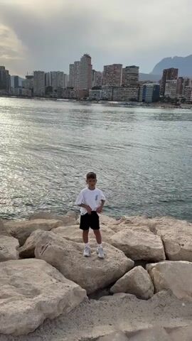 A young boy stands on a rocky shoreline. He is wearing a white t-shirt with a graphic print, black shorts, and white sneakers. He is facing the camera and making hand gestures. Behind him, a large body of water stretches out, with a city skyline visible in the distance. The sky is overcast. The boy moves his hands and poses for the camera. The boy moves his right hand up and then down and then puts his hand down. The boy then moves his left hand up and then moves his left hand down and then puts his left hand down. The boy moves his left hand up and then moves his left hand down and then puts his left hand down.