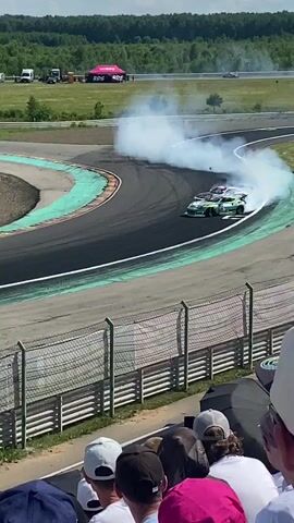 A drift car is shown maneuvering around a track. The car is predominantly white and green, and is producing a large amount of white smoke from its tires as it slides. Spectators are visible in the foreground, looking towards the track. In the background, there are trees, buildings, and other vehicles on what appears to be a race track or event venue. The weather is sunny and appears to be daytime.