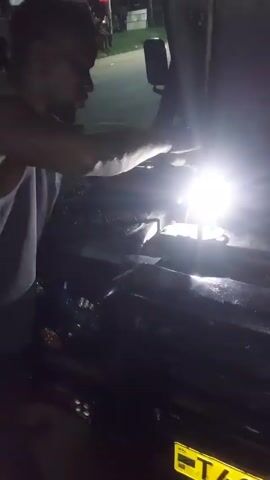 At night, a person wearing a tank top is working on a car engine. The hood is up and a bright light illuminates the engine bay. The person is reaching into the engine compartment, presumably making adjustments or repairs. The background shows a street with parked vehicles and streetlights. The scene is dimly lit, with the primary source of light coming from the work light.