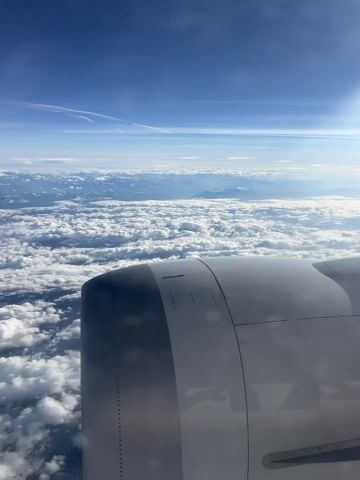 A daytime aerial shot from an airplane window captures the vast expanse of clouds and distant mountains, likely over Erkheim, Germany, given the geographical context. The bright blue sky is visible above the cloud cover, with the sun casting a soft glow. The right side of the frame is dominated by the gray and white casing of an airplane engine, which partially obscures the view of the clouds below. The engine's surface reflects some of the sunlight. Below the airplane, thick, fluffy white clouds stretch out to the horizon, creating a soft, uneven carpet. Beyond the clouds, in the distance, snow-capped mountain ranges are faintly visible through gaps and above the cloud layer. The sound of the airplane engine running is audible throughout the video, emphasizing the in-flight experience. The overall scene conveys a sense of peaceful travel high above the earth.