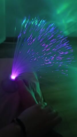 The video shows a person holding a fiber optic lamp in their hand. The lamp is on and emitting blue and green light. The person is sitting on a bed in a dimly lit room with green light. A white curtain is visible in the background. The person briefly touches the base of the lamp before moving it.