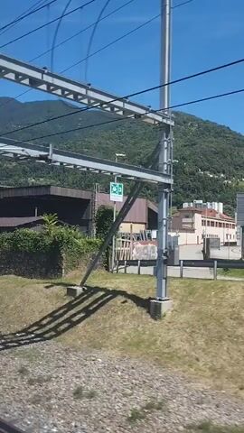 A video shows a view from a moving train passing by a landscape with buildings and mountains. The train is traveling through a rural or suburban area with green hills and trees in the background. The buildings appear to be industrial or commercial, with one prominent building displaying the text "FERRIERE CATTANEO SA". The sky is clear and blue, indicating a sunny day. The video captures the motion of the train as it moves through the scenery.