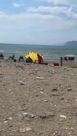 The video shows a beach scene with a sandy shoreline, a calm sea, and a partially cloudy sky. In the middle ground, two people are under a yellow and red polka-dotted umbrella. One person is sitting, and the other is standing and appears to be looking at items near the umbrella.  To the left, a person is seated in a beach chair, and to the right, two empty beach chairs are visible.  The beach is covered with a mix of sand and small stones.  In the distance, there is a landmass visible on the horizon.