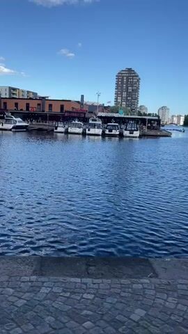 The video shows a harbor scene with several boats docked along a waterfront. The water is a deep blue, with small ripples on the surface. The foreground is a cobblestone walkway, and behind the boats are buildings, some of which appear to be restaurants or shops with outdoor seating. Further in the background, a tall, modern building with a dark facade stands out, along with other contemporary architecture. The sky is blue with scattered white clouds, suggesting a pleasant, possibly sunny day. The time appears to be daytime, likely late afternoon or early evening given the angle of the light. There are people visible on the docks and at the outdoor seating areas of the establishments, indicating a lively atmosphere. The scene evokes a sense of calm and leisure, typical of a waterfront setting. No discernible text is clearly visible in the video, except possibly for some signage on the buildings which are too indistinct to read. The overall impression is one of a modern European city's harbor area, likely in a place like Karlstad, Sweden, given the architectural style and waterfront development.